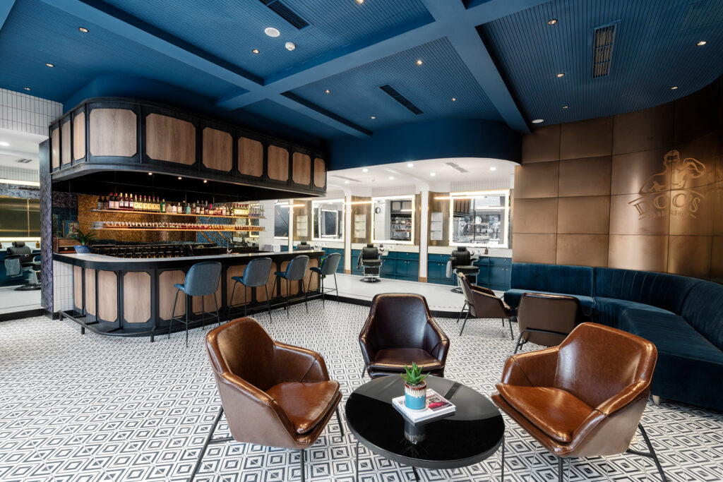 blue, brown & white colored barbershop with small brown chairs & wood-framed bar in center of the room. A blue velvet sofa off to the right 